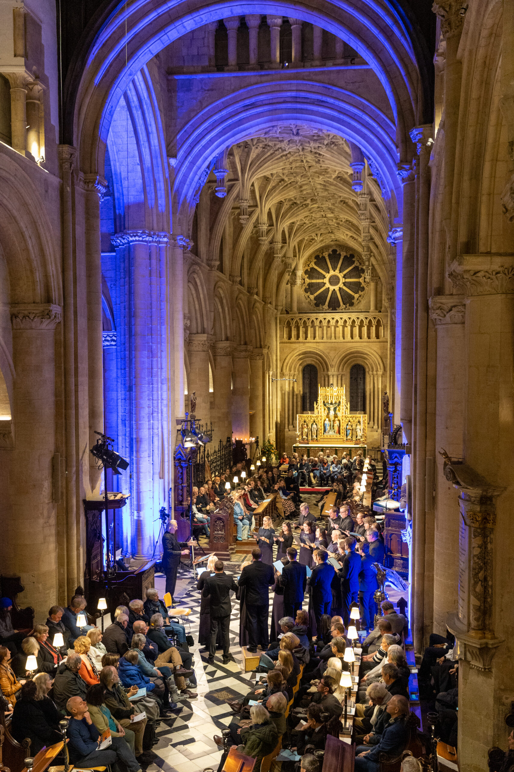 A shot over rows of audience with a choir singing in a blue lit, high ceilinged old stone building.