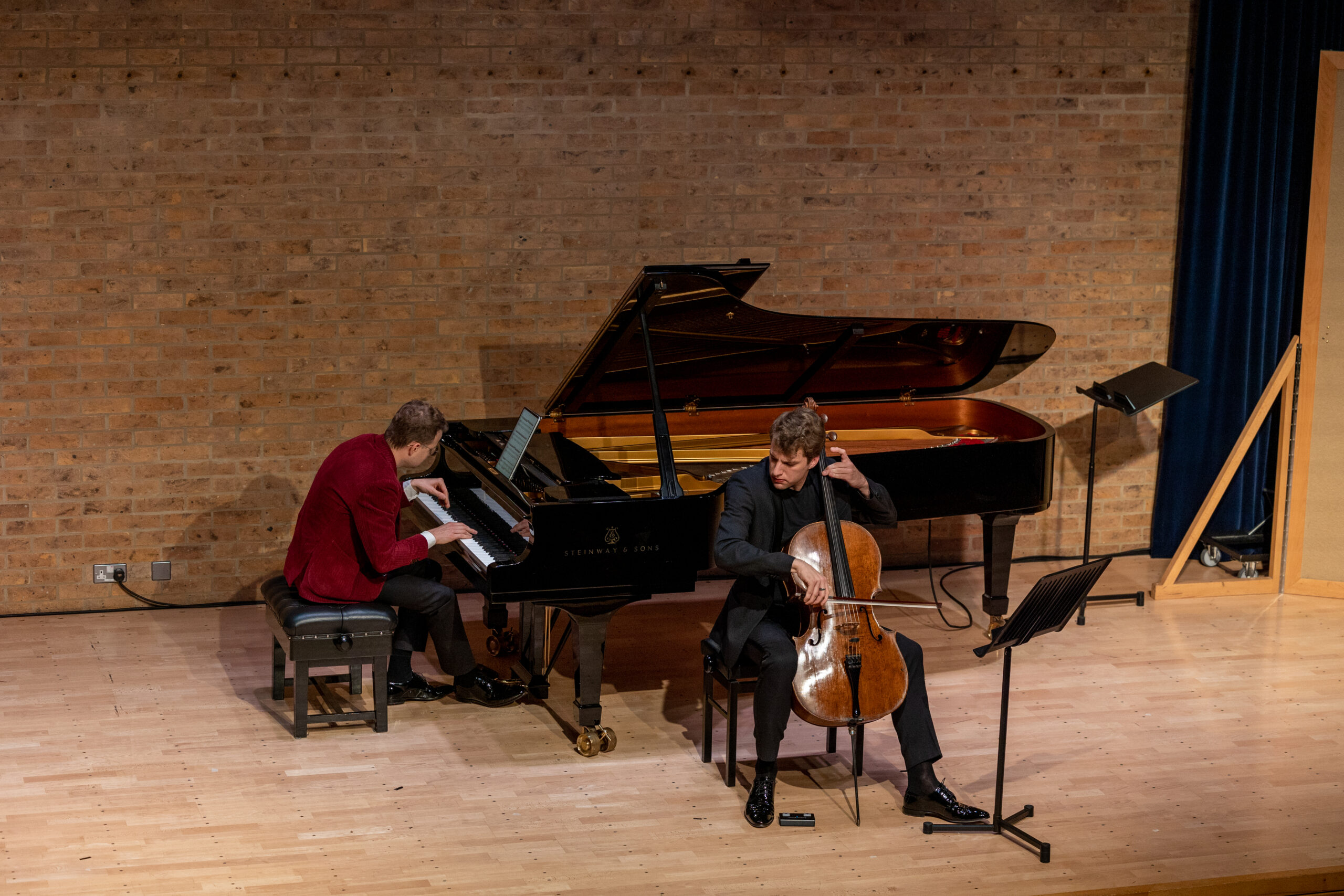 Man in red jacket playing piano on stage with another man playing the cello sitting in front of the piano.
