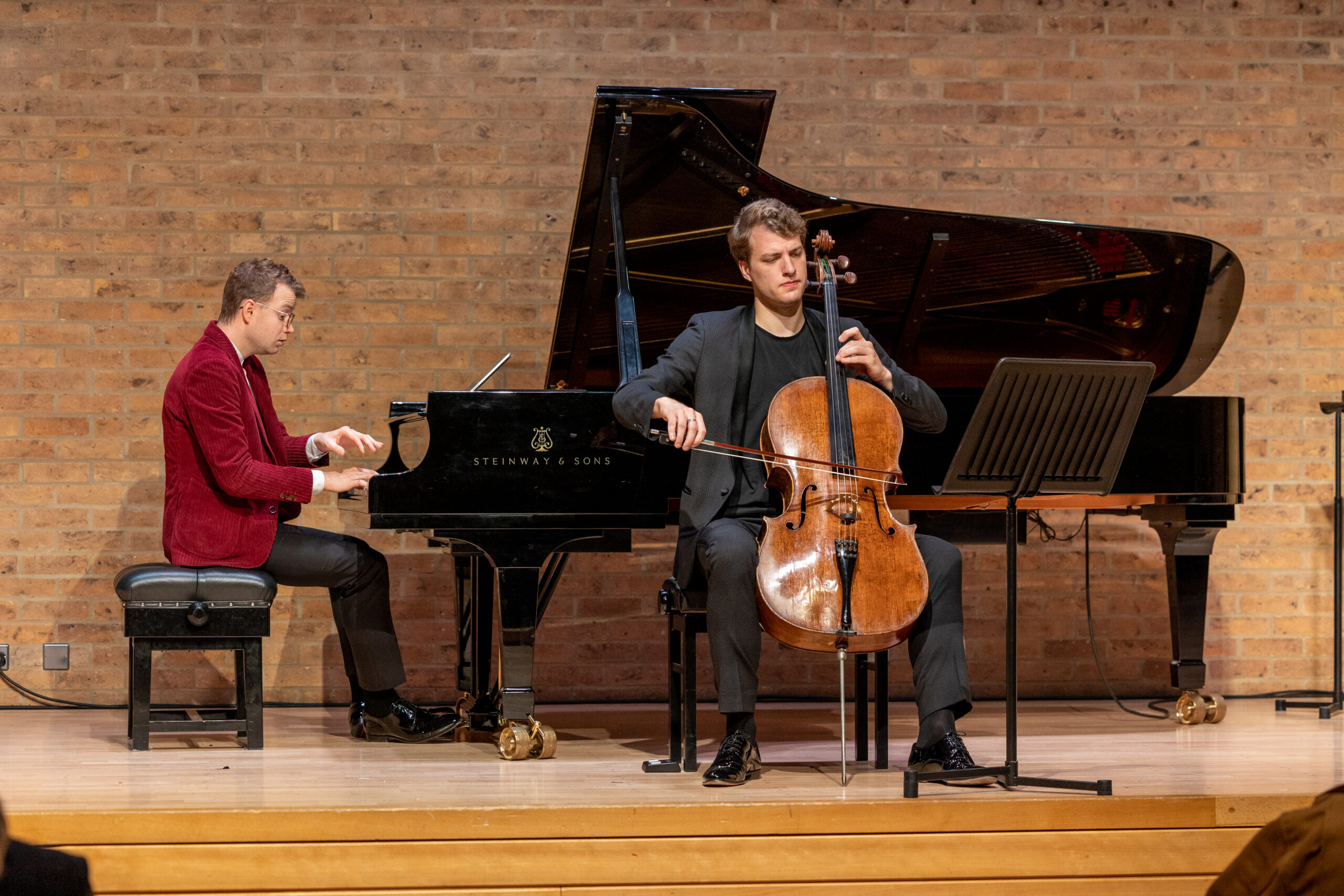 Close up of Man in red jacket playing piano on stage with another man playing the cello sitting in front of the piano.
