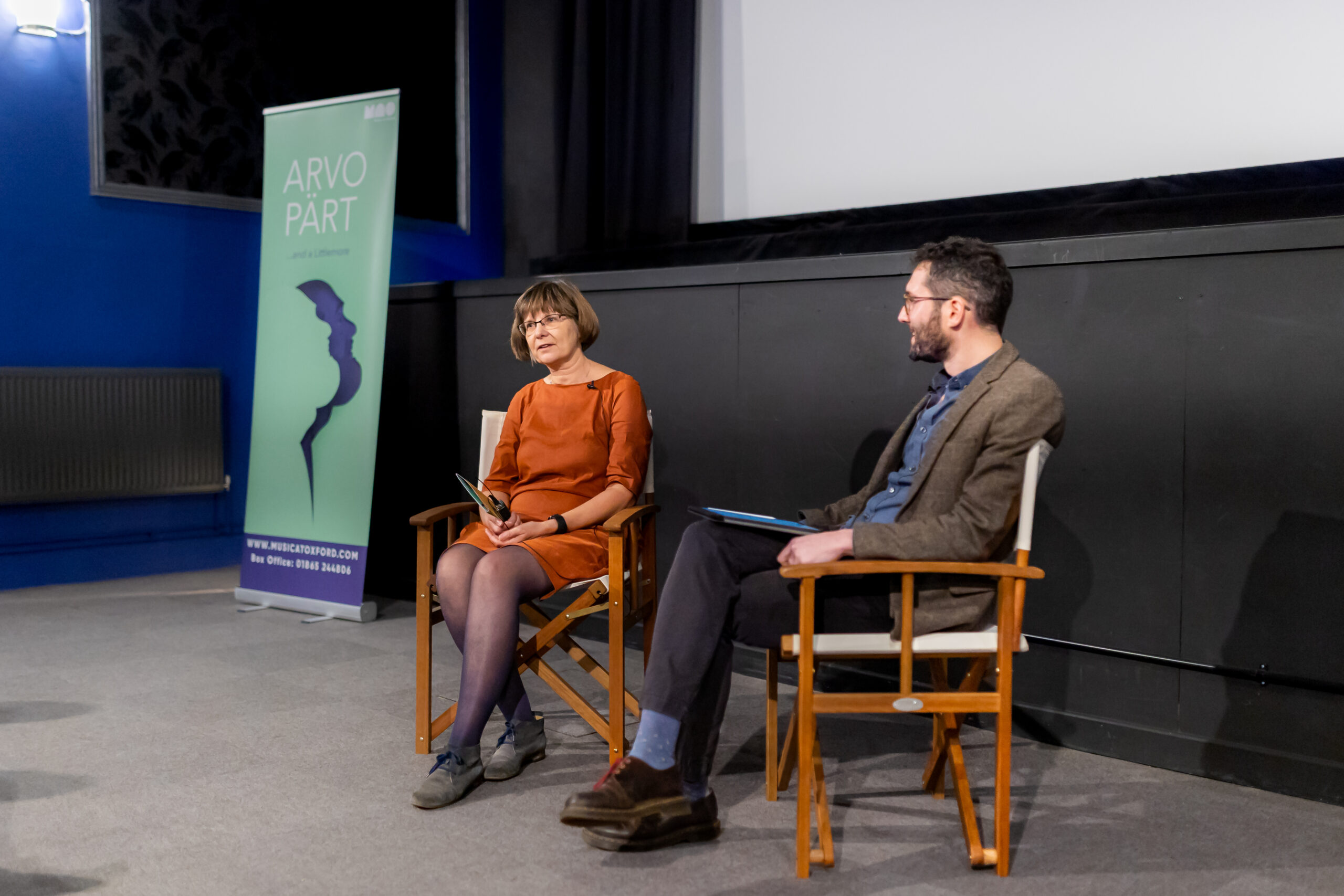 A man and a women sit in chairs below a screen and talk. A green ARVO PÄRT...and a Littlemore festival banner is on the left next to the woman.