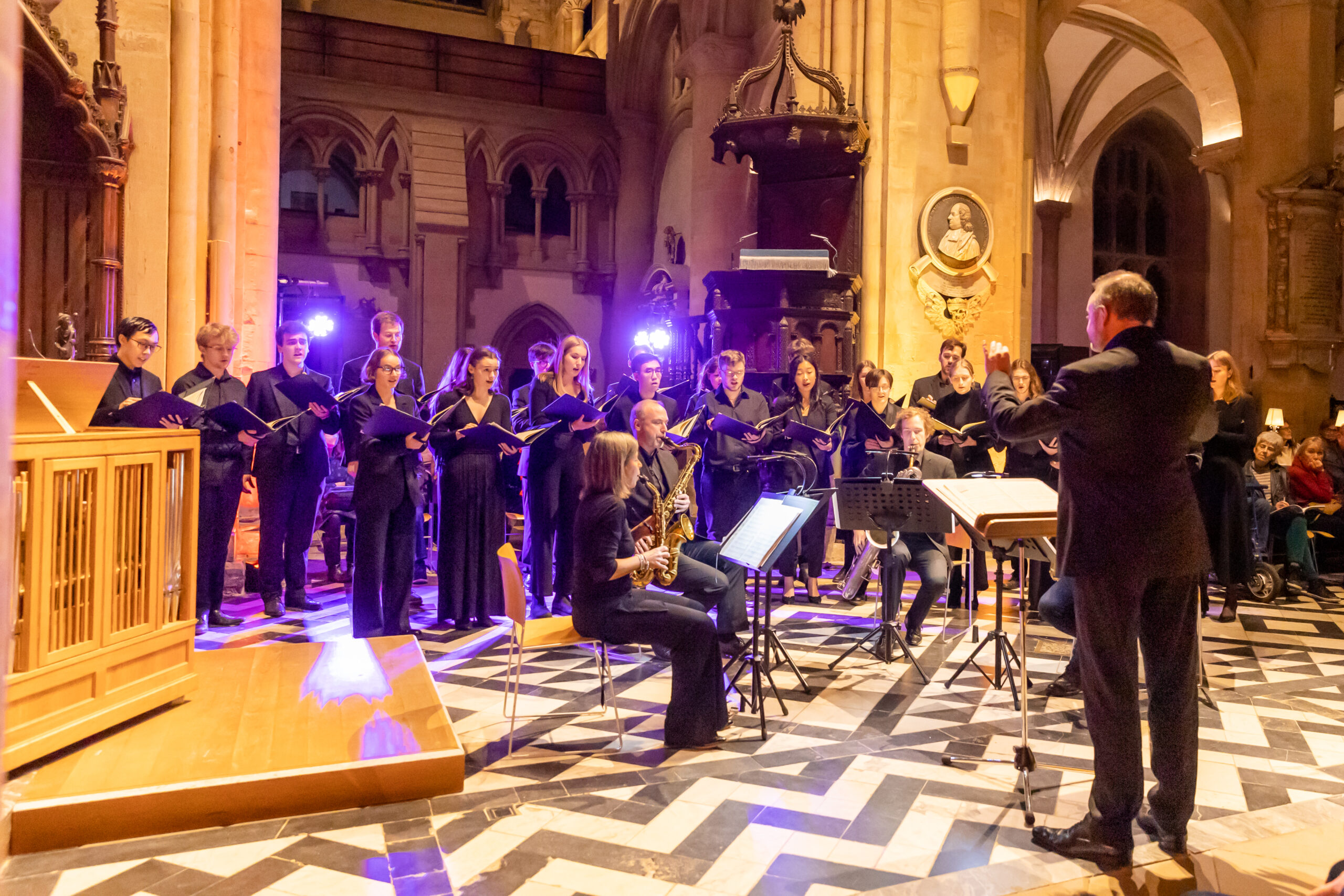 A choir with a saxophone quartet sitting in front and a conductor perform. It's an old building with a black and white zig-zag floor and lit with pink lighting.