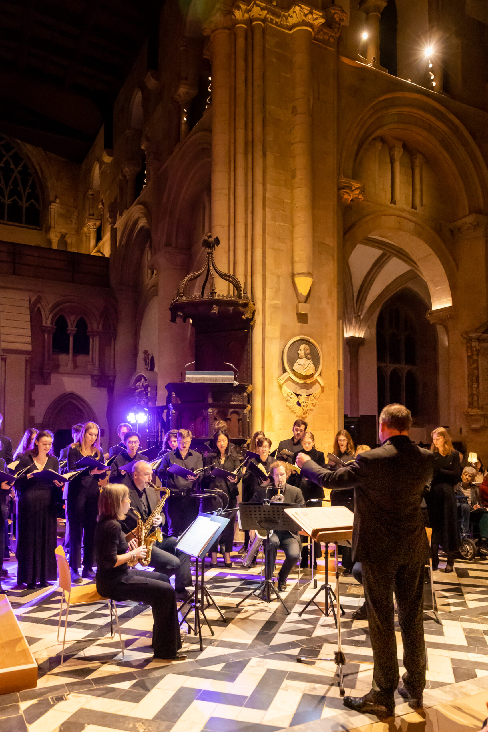 A choir with a saxophone quartet sitting in front and a conductor perform. It's an old building with a black and white zig-zag floor and lit with pink lighting.