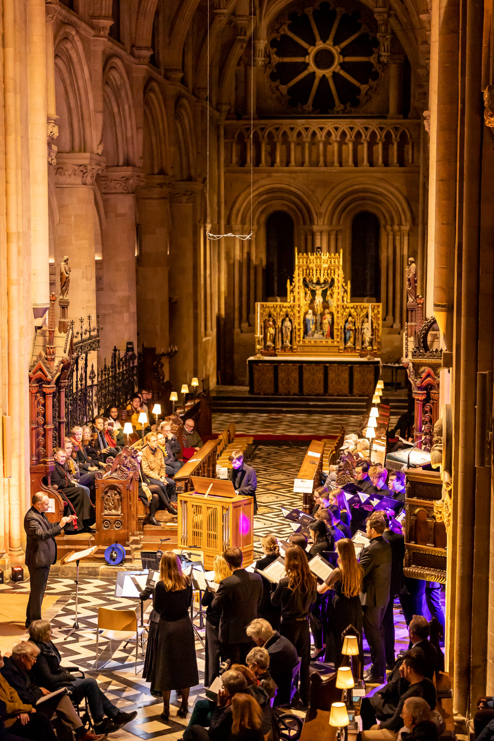 A choir and organ performing with their conductor in the crossing of the cathedral. Audience members can be see sitting to the left and right of the choir with the alter at the back of the photo.