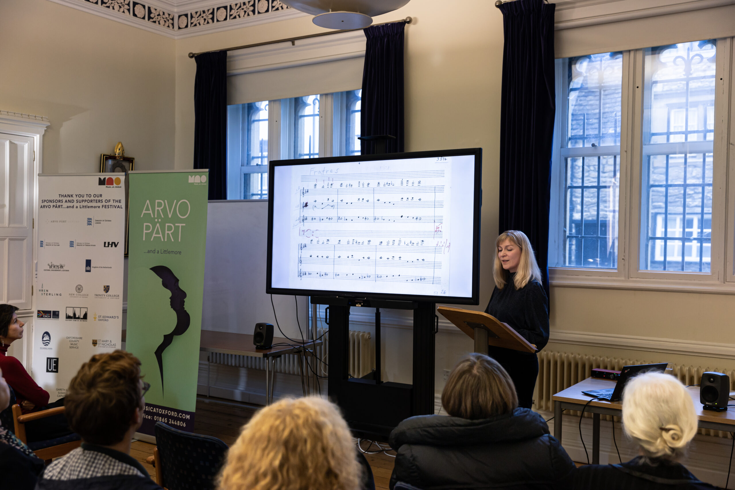 A woman stands behind a lectern and next to a screen showing a musical composition manuscript. Some audience members are visible in the foreground. To the left of the image is a green ARVO PÄRT...and a Littlemore festival banner.