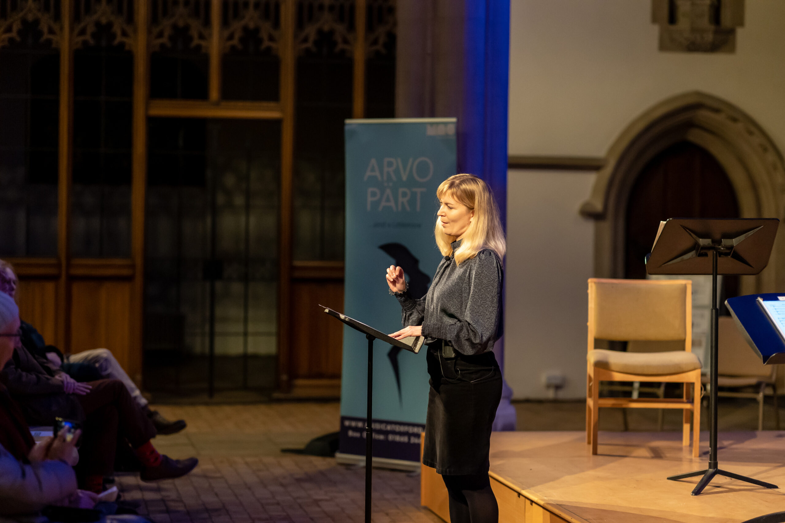 A side-on view of a woman standing behind a lectern in front of a stage. Some audience members are visible in the foreground. To the right of the woman is a blue ARVO PÄRT...and a Littlemore festival banner.