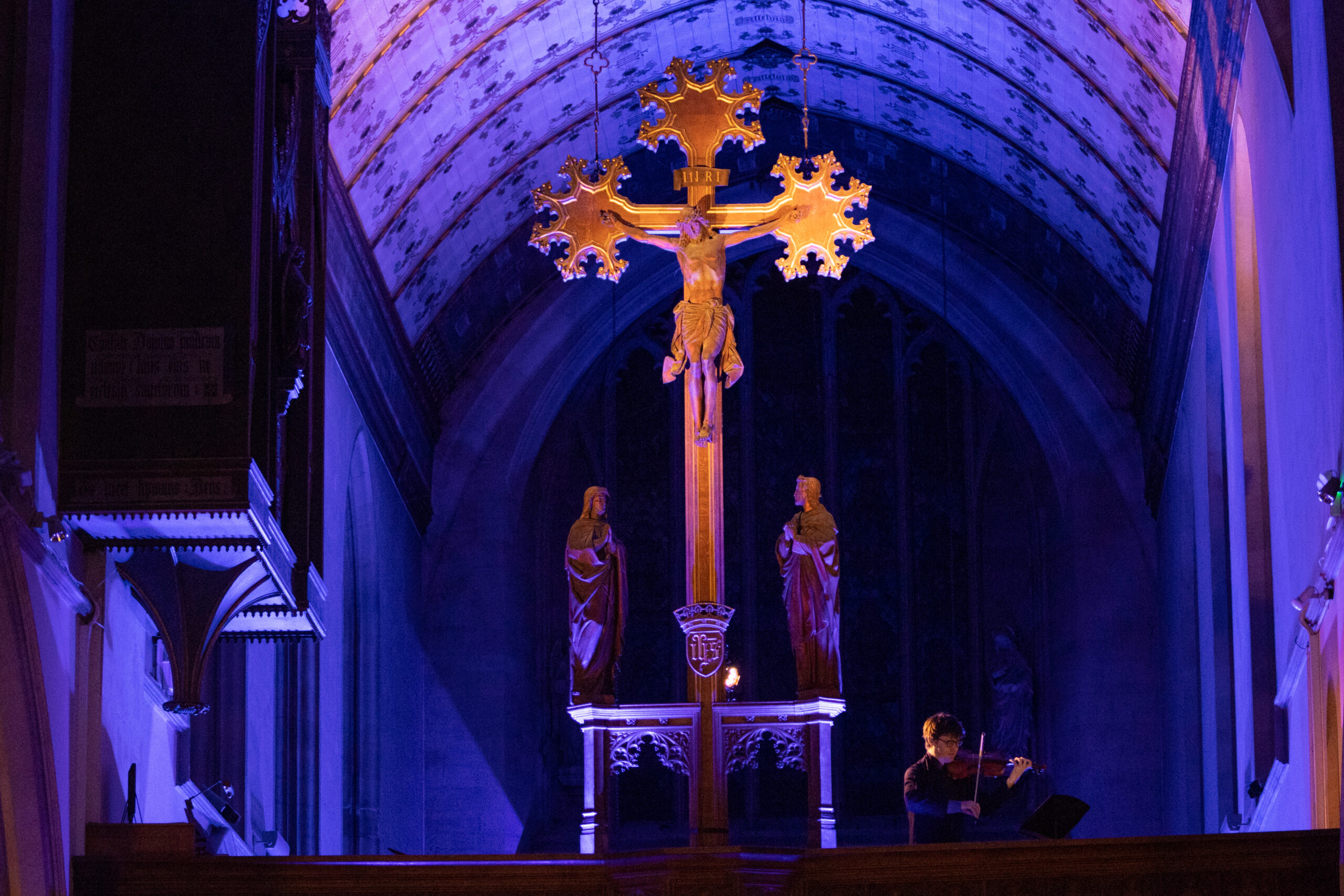 A distant shot of a man playing the violin next to a large cross on the balcony of a church. It is lit with a soft blue light.