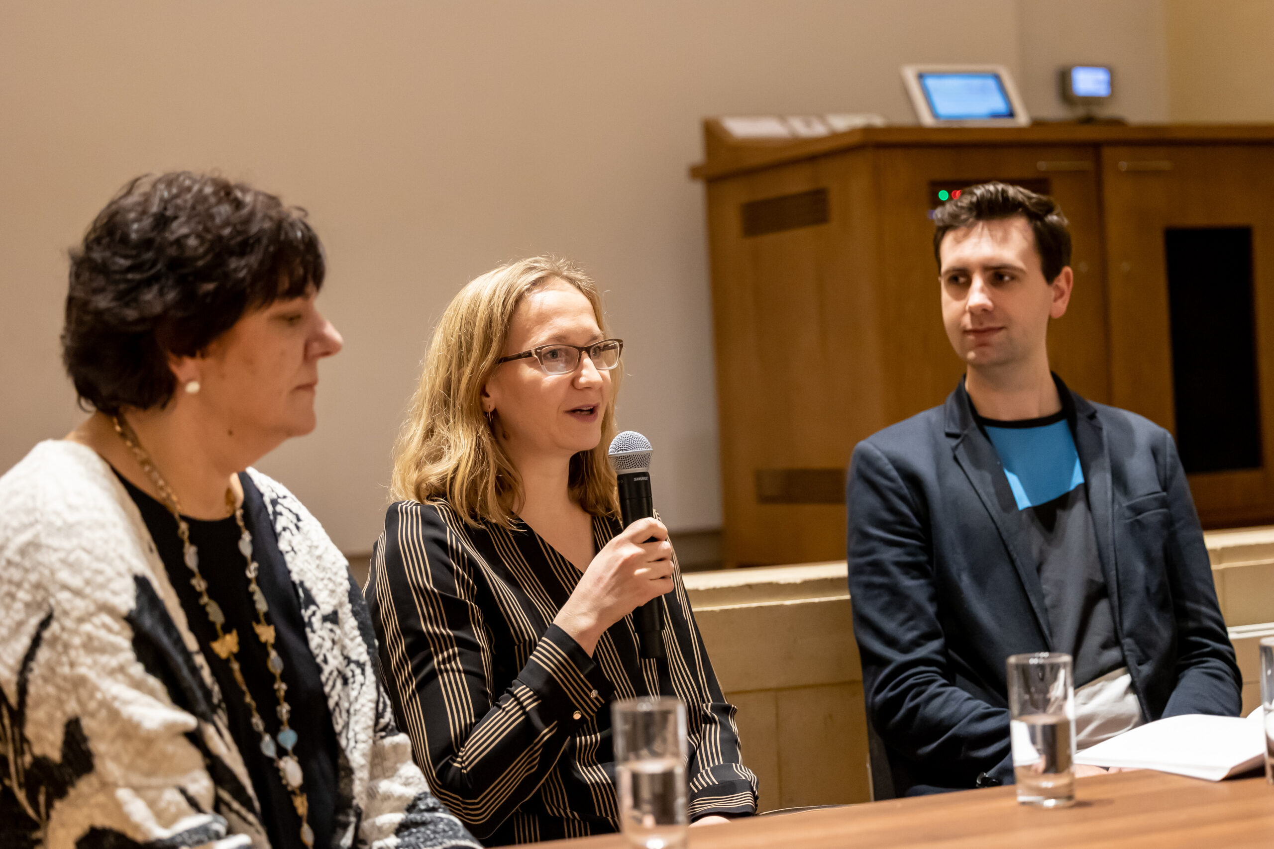 Two women and a man sit behind a table, the woman in the middle is talking into the microphone.