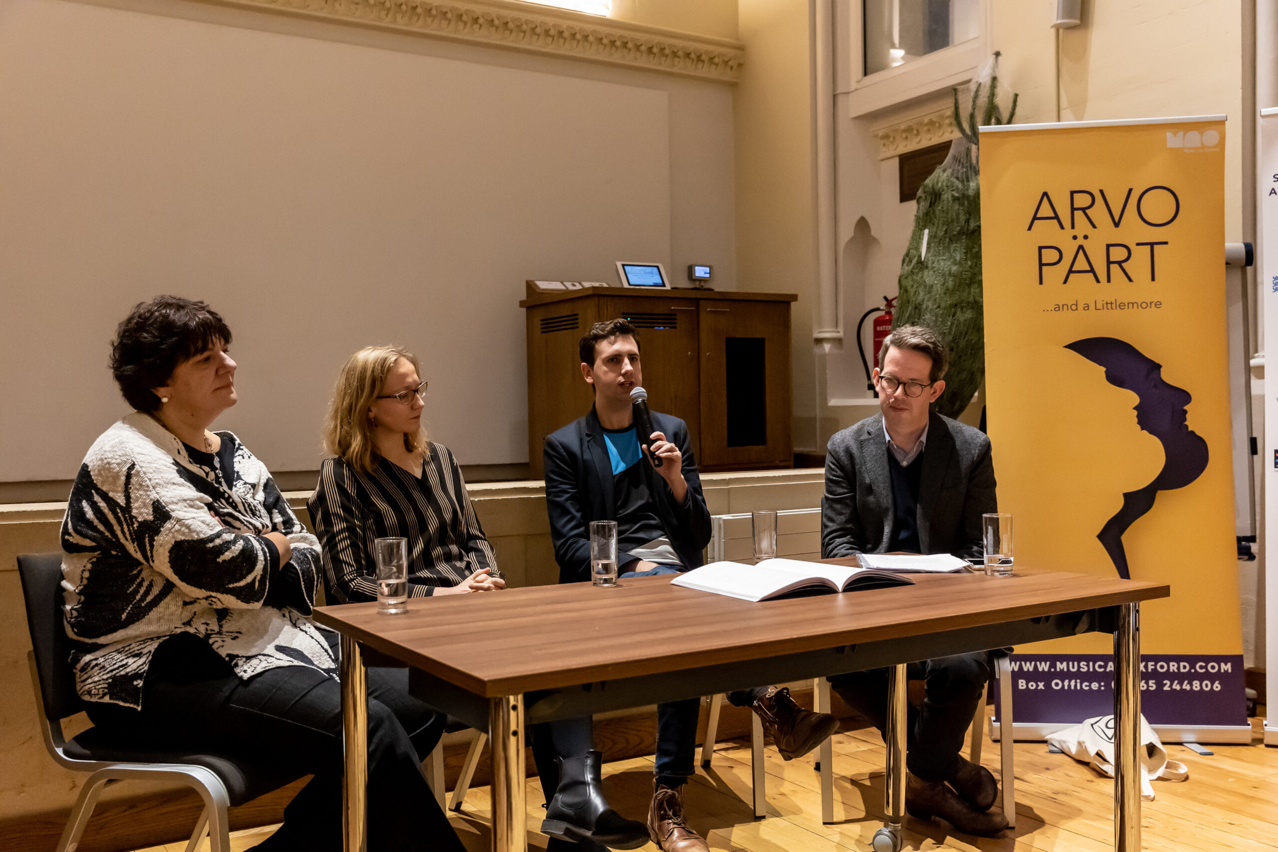 Two women and two men sit behind a desk. One of the men is talking into the microphone. A yellow ARVO PÄRT...and a Littlemore festival banner is behind them to the right of the shot.