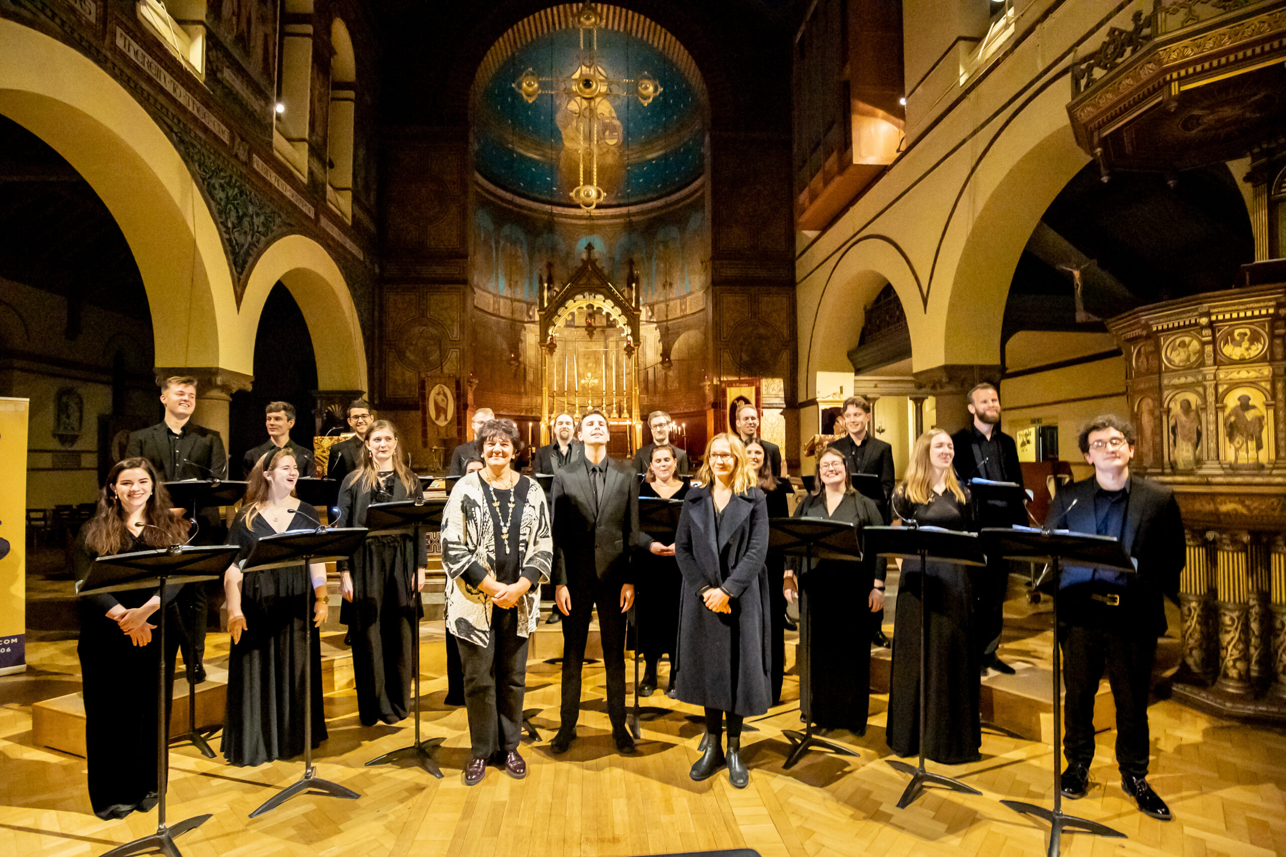 A choir, their director and two women composers stand in front of the alter of a decorated church.