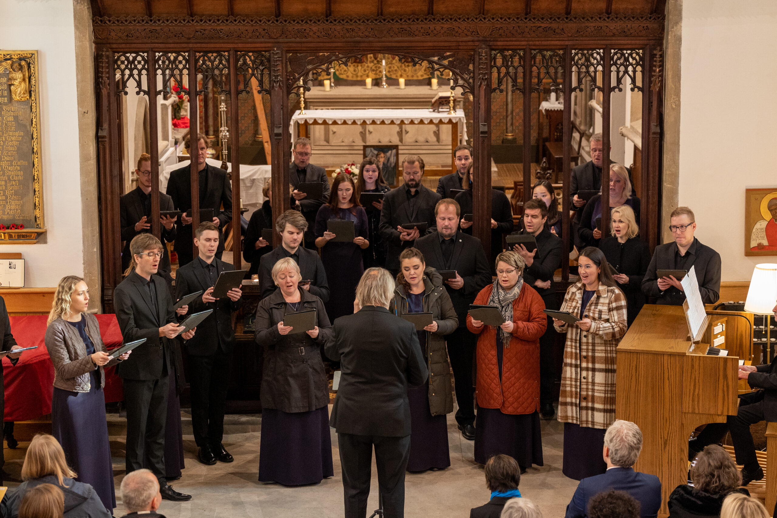 A choir sing in coats in a church for an audience of which you can see a few heads at the bottom of the shot. Their director stands in the middle facing the choir.