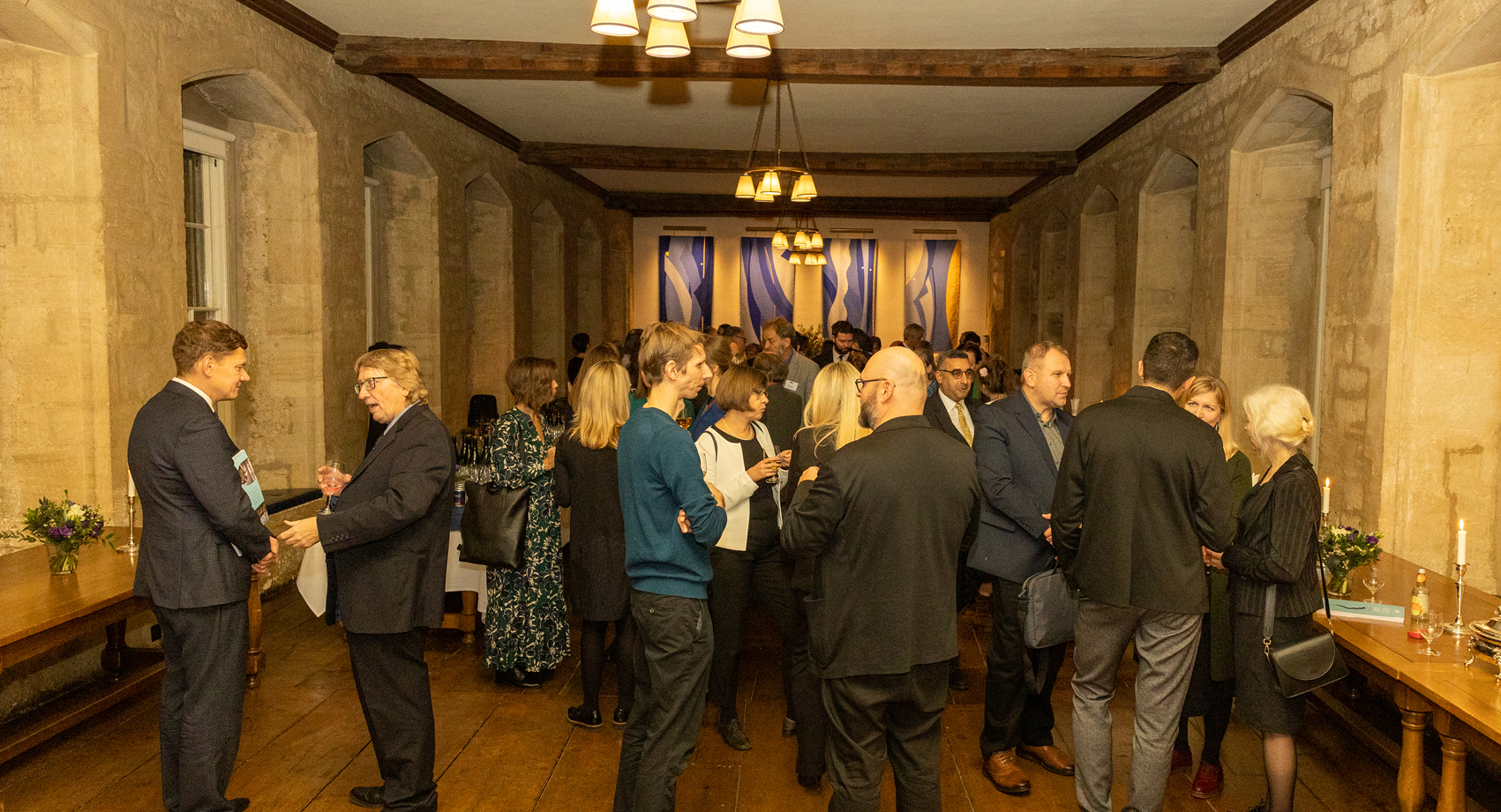 Groups of people gather, talk and drink at an event in a low ceilinged, stone room.