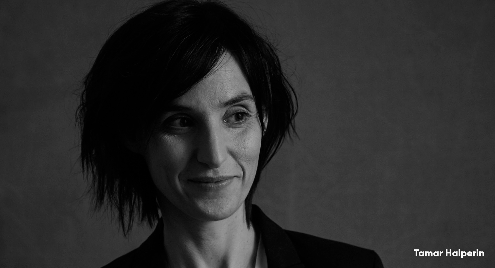 A black and white headshot of Tamar Halperin smiling with short hair, looking to the side, away from the camera.